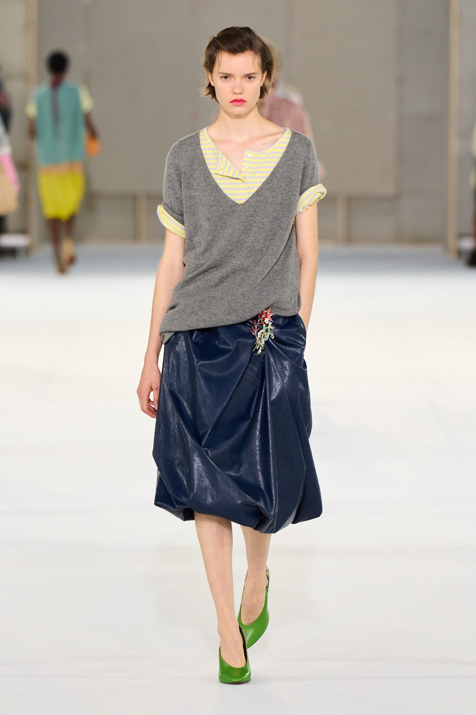 A model wearing a colorful jeweled floral brooch fastened at the waist of a navy skirt during a September 30, 2026 Paris Fashion Week runway show