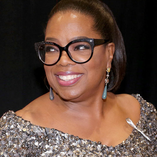 Oprah Winfrey smiling in bold black eyeglasses and dangling gemstone earrings at an event; celebrity style example, not Suzy Levian jewelry