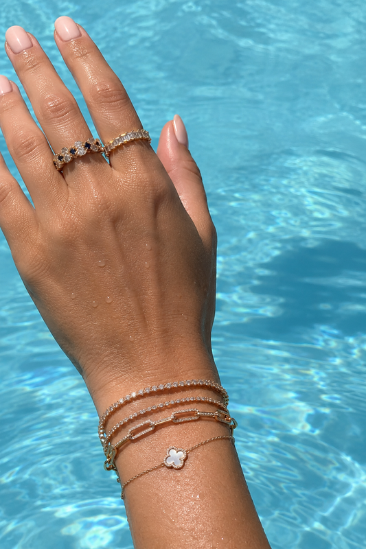 Hand wearing Suzy Levian-style rings and stacked bracelets over bright blue pool water for a summer jewelry care guide