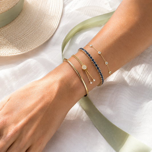 Summer bracelet stack with gold bangle, blue gemstone bracelet, and delicate station bracelets on white linen